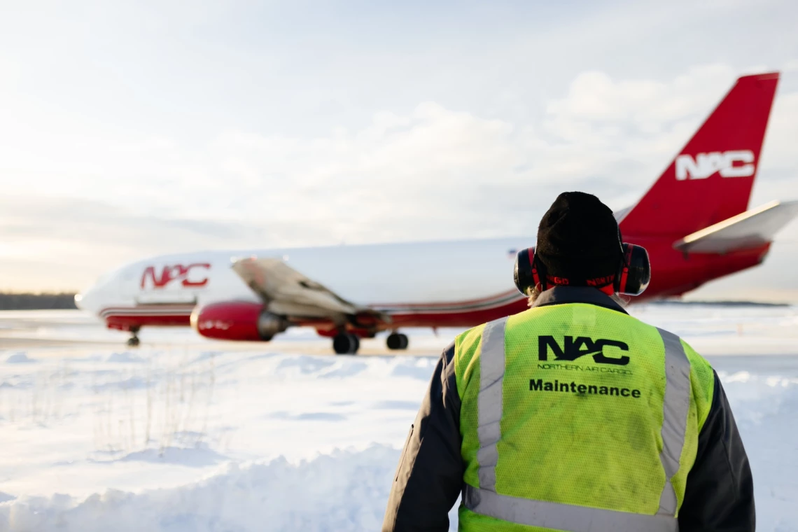 A Northern Air Cargo maintenance employee standing in the snow watching a plane take off.