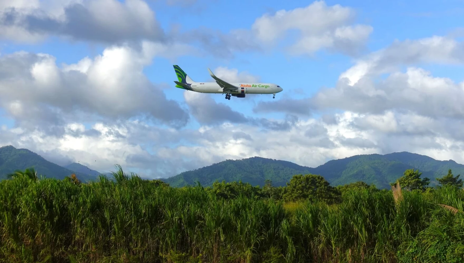 An Aloha Air Cargo plane in the sky with a lush, tropical trees and mountains underneath.