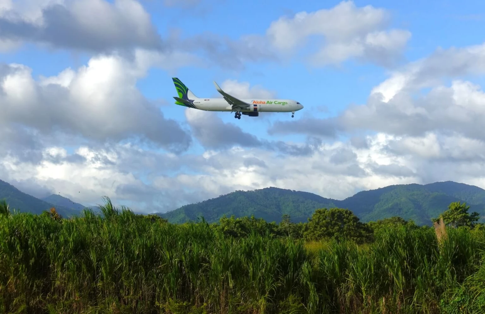 An Aloha Air Cargo plane in the sky with a lush, tropical trees and mountains underneath.