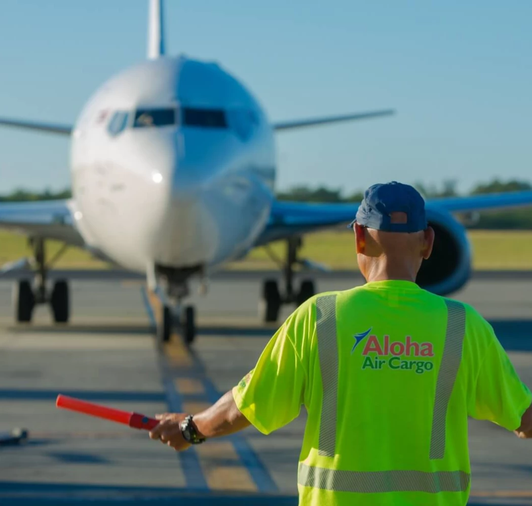 An Aloha Air Cargo employee using orange safety batons to help signal a plane on the runway.