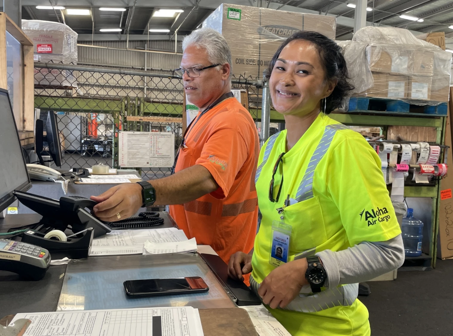 Aloha Air Cargo employees standing at the customer service desk, smiling at the camera and answering the phone.