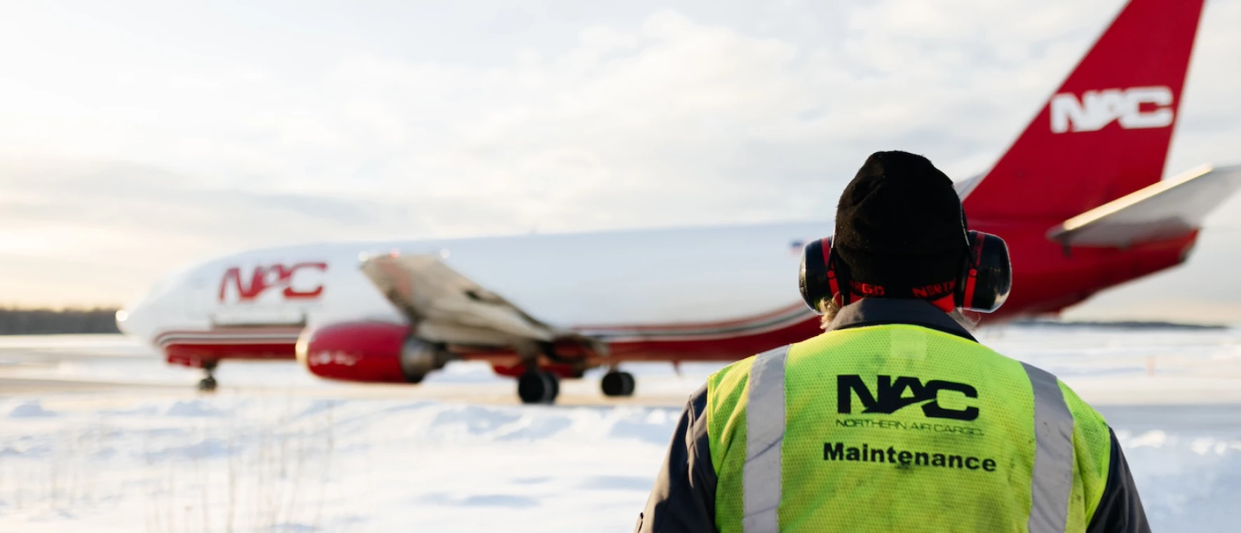 A Northern Air Cargo maintenance employee standing in the snow watching a plane take off.